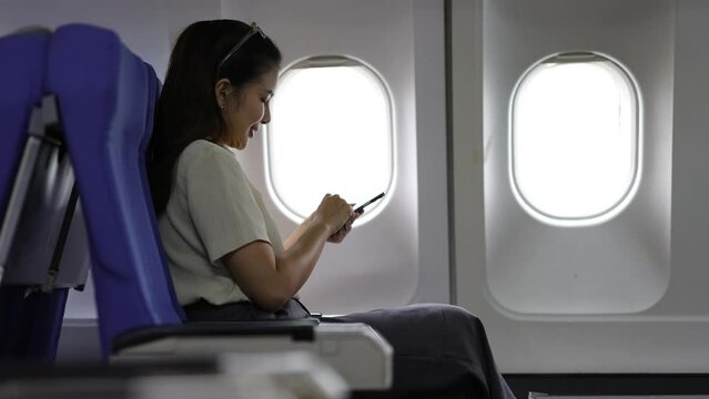 Happy young Asian woman traveling alone playing with mobile phone on airplane, she is texting with friends.