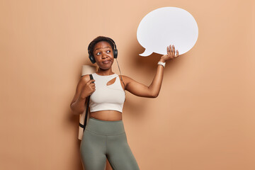 Horizontal shot of thoughtful African woman dressed in sportswear listens music via headphones carries rolled karemat holds communication bubble for your promotion isolated over brown studio wall