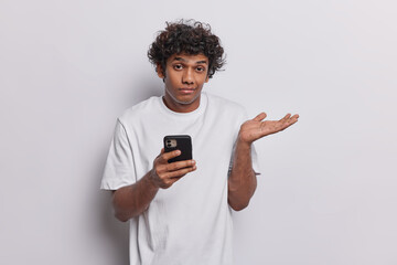 People emotions concept. Indoor photo of young confused Hindu man standing isolated in centre on white background holding smartphone can't find necessary information on internet wearing t shirt
