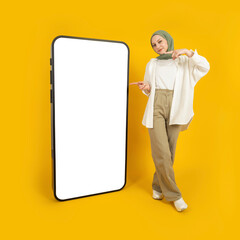 Pointing finger on big mobile phone, full body young caucasian smiling muslim woman in hijab standing near huge smartphone with empty blank screen mock up. Recommending new application concept idea.