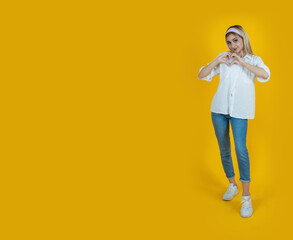 Full body blonde girl holding hands over chest in heart shape gesture. Standing over isolated yellow studio background. Love concept. Copy space. Advertising graphic element design.