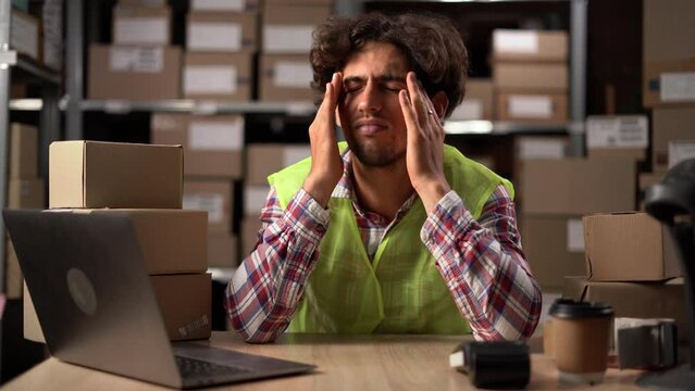 Tired young man fatigued from computer work, stressed man suffer from headache sit at warehouse using laptop