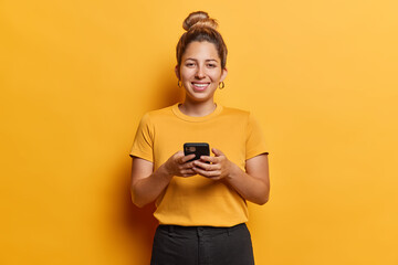 Cheerful attractive young woman with hair bun holds mobile phone reading comments in social networks smiles happily being in good mood wears casual t shirt black trousers isolated on yellow background - obrazy, fototapety, plakaty