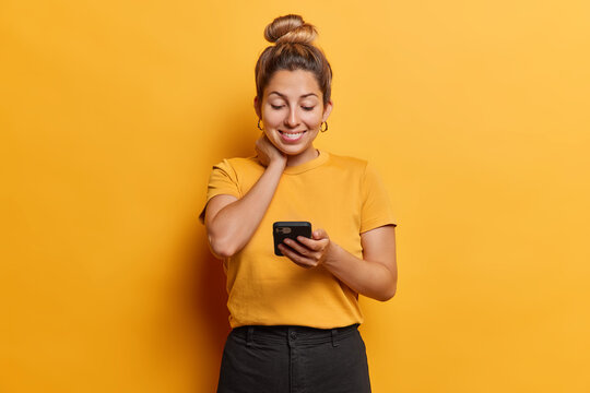 Waist up shot of pleased European woman smiles gently keeps hand on neck focused in smartphone screen enjoys online communication wears casual t shirt and black trousers isolated on yellow background
