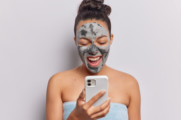 Indoor shot of emotional Latin woman with facial clay mask on face exclaims loudly holds mobile...