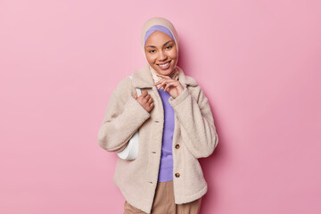 Studio shot of pretty tender Muslim woman wearing hijab and fur coat poses with small bag on...