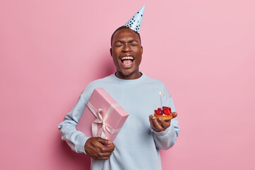 Studio shot of young happy excited smiling broadly African american boy celebrating birthday holding present and cake with candle standing isolated in centre on pink background wearing hat and sweater