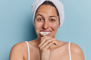 Indoor shot of young European woman brushes teeth with toothbrush stands bare shouldered against blue background wears bath towel on head casual t shirt concentrated at camera. Hygiene concept