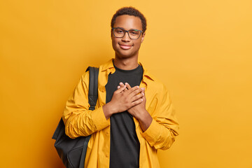 People positive emotions concept. Studio photo of young glad smiling African american male student with standing in centre isolated on yellow background showing love keeping hands near heart