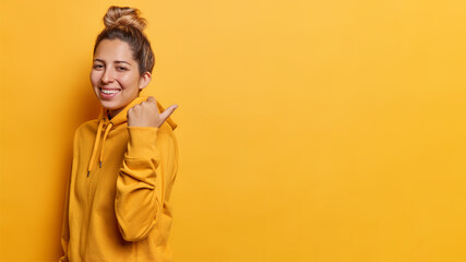 Horizontal shot of young European woman with hair bun smiles pleasantly points thumb away shows awesome promo dressed in casual hoodie isolated over yellow background. Follow this way please