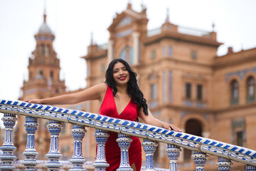 Young and beautiful brunette and latin woman, dressed in short red dress and red shoes visits the famous square in seville, spain. Woman leaning on the railing of a tiled bridge and is happy. Travel.