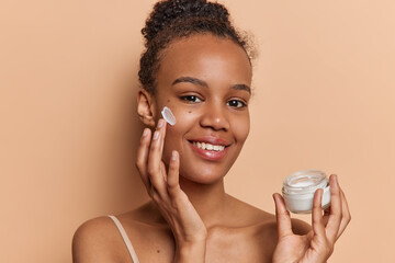 Beauty and cosmetology concept. Indoor photo of young pretty happy smiling African american lady standing in centre isolated on beige background holding applying cream under eyes to reduce wrinkles