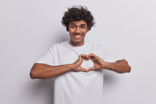 Romance concept. Positive handsome curly haired Hindu man shapes heart gesture with hands dressed in casual t shirt smiles pleasantly isolated over white background says I love you. Body language