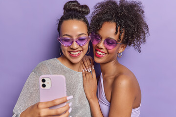 Cheerful brunette women take selfie via smartphone click photos on front camera wear sunglasses and stylish clothing isolated over purple wall spending time together filming content for her video blog - Powered by Adobe