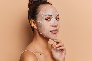 Sideways shot of dark haired Latin woman with beauty sheet mask on face for rejuvenation keeps hand...
