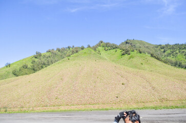 Lush green mountain slopes meet a dry, tan-colored hillside, with a paved road in the foreground.