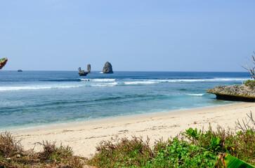 Fototapeta premium Coastal scene with a sandy beach, ocean waves, and rock formations under a blue sky.