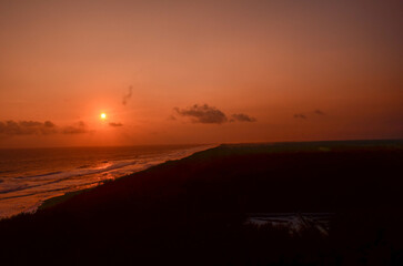 Sunset over the ocean, casting a warm orange glow across the sky and reflecting on the water along the coastline.