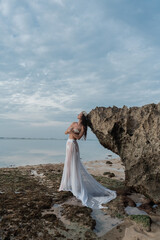 Fototapeta na wymiar A girl with a veil poses in a crown of shells against the backdrop of a reef and rocks