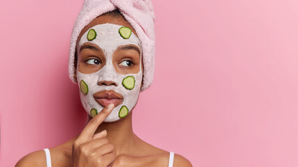 Beauty and cosmetology. Studio shot of young dreamy relaxed European girl applying clay face mask and slices of cucumber standing isolated on left on pink background looking at space for promotion - Powered by Adobe