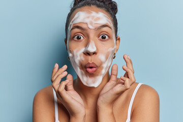 Horizontal shot of impressed Latin woman washes face with white soap cleans pores keeps lips folded feels stunned isolated over blue background. Skin care pampering and daily hygiene concept