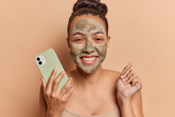 Self care and beauty concept. Positive Latin woman makes beauty treatments after bath has face covered with clay moisturising mask uses mobile phone clenches fist isolated over brown background