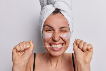 Beautiful positive young woman uses dental floss for cleaning teeth applies beauty patches under eyes to reduce wrinkles wears bath towel on head and black t shirt isolated over white background