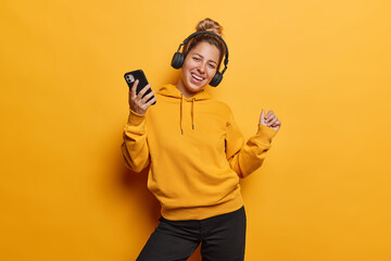 People positive emotions concept. Indoor photo of young happy smiling excited woman listening to music on headphones dancing holding smartphone wearing casual clothes isolated on yellow background