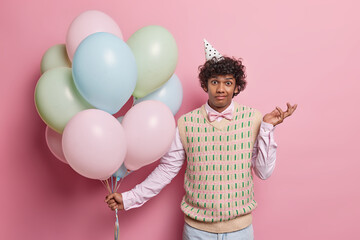 Hesitant birthday man shrugs shoulders with clueless expression holds bunch of inflated colorful balloons doesnt know why guests didnt come isolated over pink background. Celebration concept - obrazy, fototapety, plakaty