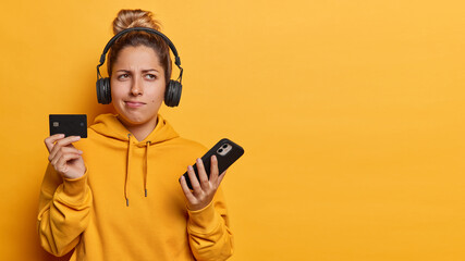 People emotions concept. Indoor photo of young pretty dreamy European girl keeping headphones on head listening to music holding smartphone and card standing on left isolated on yellow background