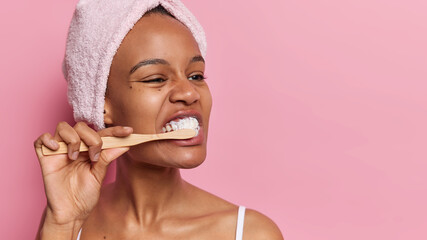 Beauty procedures. Studio shot of young beautiful European lady after shower or bath with towel on head cleaning teeth as part of daily routine standing on left isolated on pink background with space
