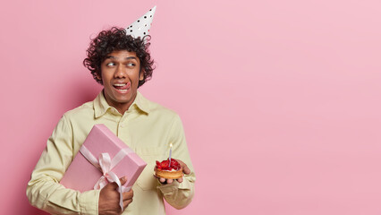 Dreamy thoughtful Hindu man with curly hair holds present box and cupcake gets congratulations on...