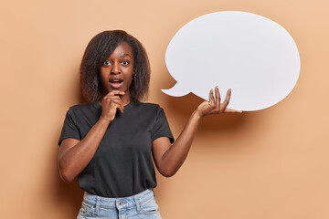 Photo of impressed dark skinned young woman with bushy hair keeps mouth opened holds blank speech bubble for your advertising content wears black t shirt and jeans isolated over brown background