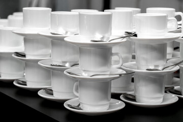 White coffee cups with saucers and spoons are stacked on a black table. Preparing for the buffet