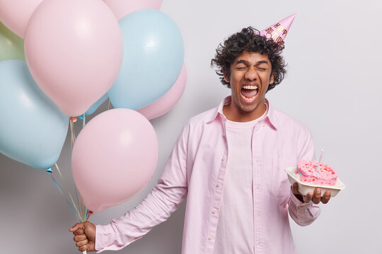 Positive Young Hindu Man Holds Bunch Of Inflated Balloons And Creamy Cake With Burning Candle Wears Festive Clothing And Party Hat Celebrates One Year Of Work Poses Against White Background.