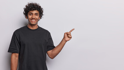 Waist up shot of Hindu curly haired man pointing at copy space for your text or advertisement shows place for promotional product presents something wears casual black t shirt isolated over white wall