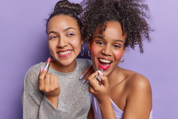 Photo of two women apply lipstick smile broadly dressed in fashionable clothes make makeup get...