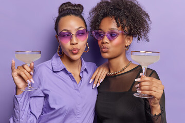 Two fashionable female friends wear stylish sunglasses and clothing hold glass of cocktails fold lips look at each other enjoy party time isolated on purple background. People and celebration concept