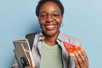 Horizontal shot of cheerful African girl with short dark hair smiles plesantly holds piece of pizza...