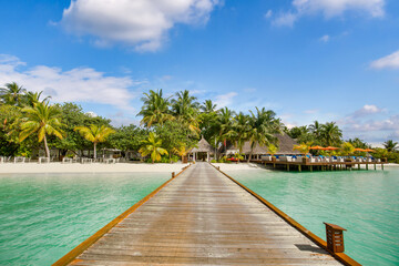 Luxury travel landscape. Water villas, wooden pier bridge leads to palm trees over white sandy shore close to blue sea, seascape. Summer panoramic vacation, beach resort on tropical island paradise
