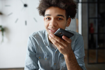 Indoor portrait of young african american man recording audio message chatting with friend in messenger on phone. Afro man in shirt dictating commands and recording notes on smartphone voice recorder