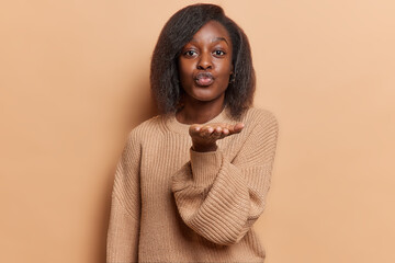 Photo of good looking dark skinned African woman sends sweet air kiss keeps palm outstreched near...
