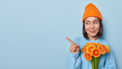Studio shot of pretty young Asian woman points index finger on blank space holds bouquet of orange daisies celebrates birthday wears orange hat and knitted jumper isolated over blue background