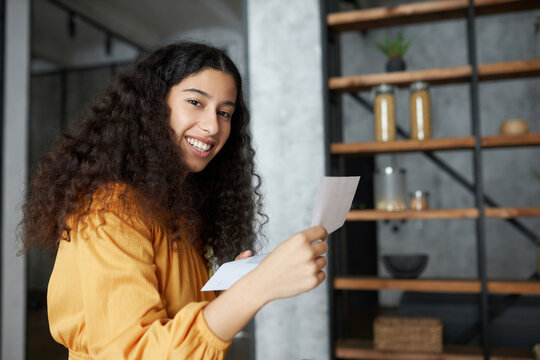 Side view indoor portrait of happy smiling african american female student holding letter with job offer, scholarship admission, money refund or loan approval standing in loft-style kitchen