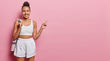 Horizontal shot of satisfied Latin woman dressed in white cropped top and shorts carries rolled...