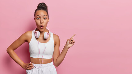 Waist up shot of impressed Latin woman dressed in white tracksuit points index finger on copy space demonstrates something shocking leads sporty lifestyle suggests to write your promotion here