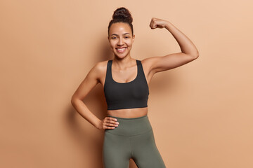 Horizontal shot of active sporty woman dressed in black cropped top and leggings shows muscles how strong she is smiles gladfully poses against brown background brags about results in sport.