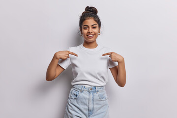 Horizontal shot of pretty dark haired woman with hair bun points index fingers on blank space of t...