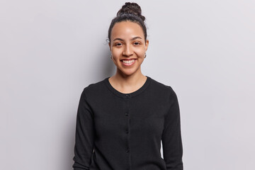 Waist up shot of cheerful Latin woman smiles gently looks directly at camera dressed in casual black jumper isolated over white background. Beautiful young smiling female model being in good mood