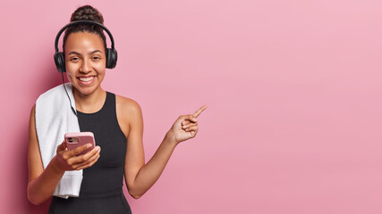 Waist up shot of Latin sportswoman dressed in black tracksuit has towel on shoulder uses mobile phone and headphones for listening music points aside isolated over pink background. Sport advertisement